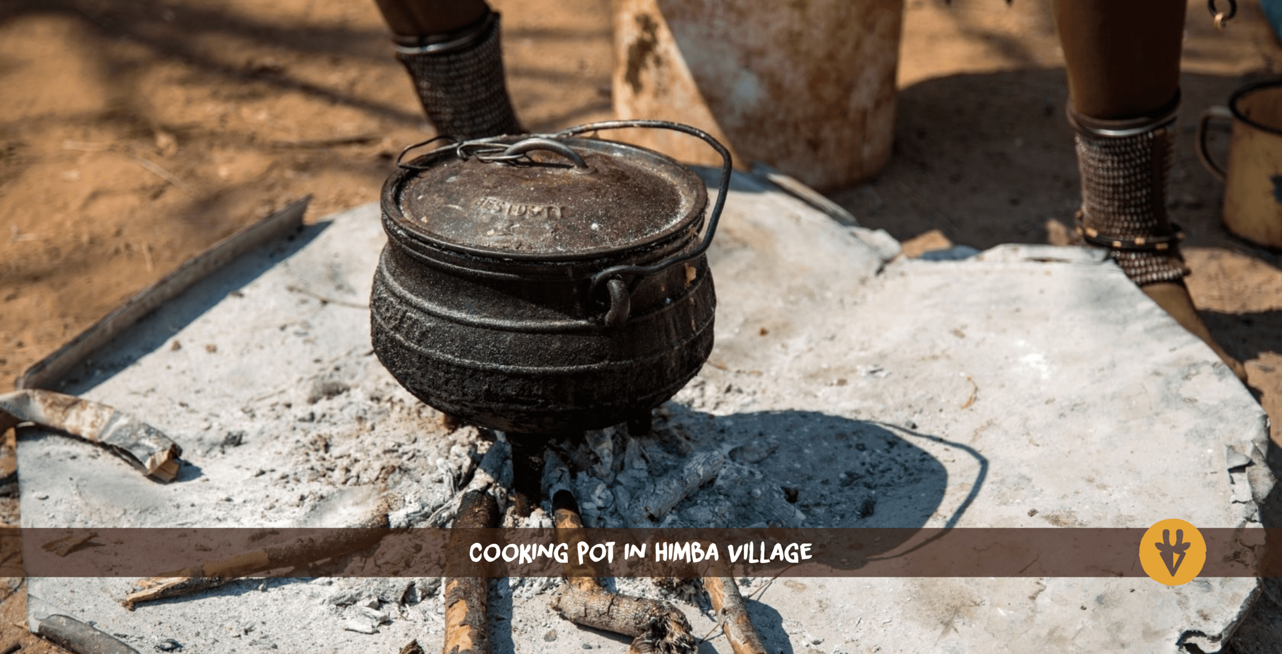 A dark-brown pot with a lid and handle standing on ash and some small branches of a former fire in a Himba village