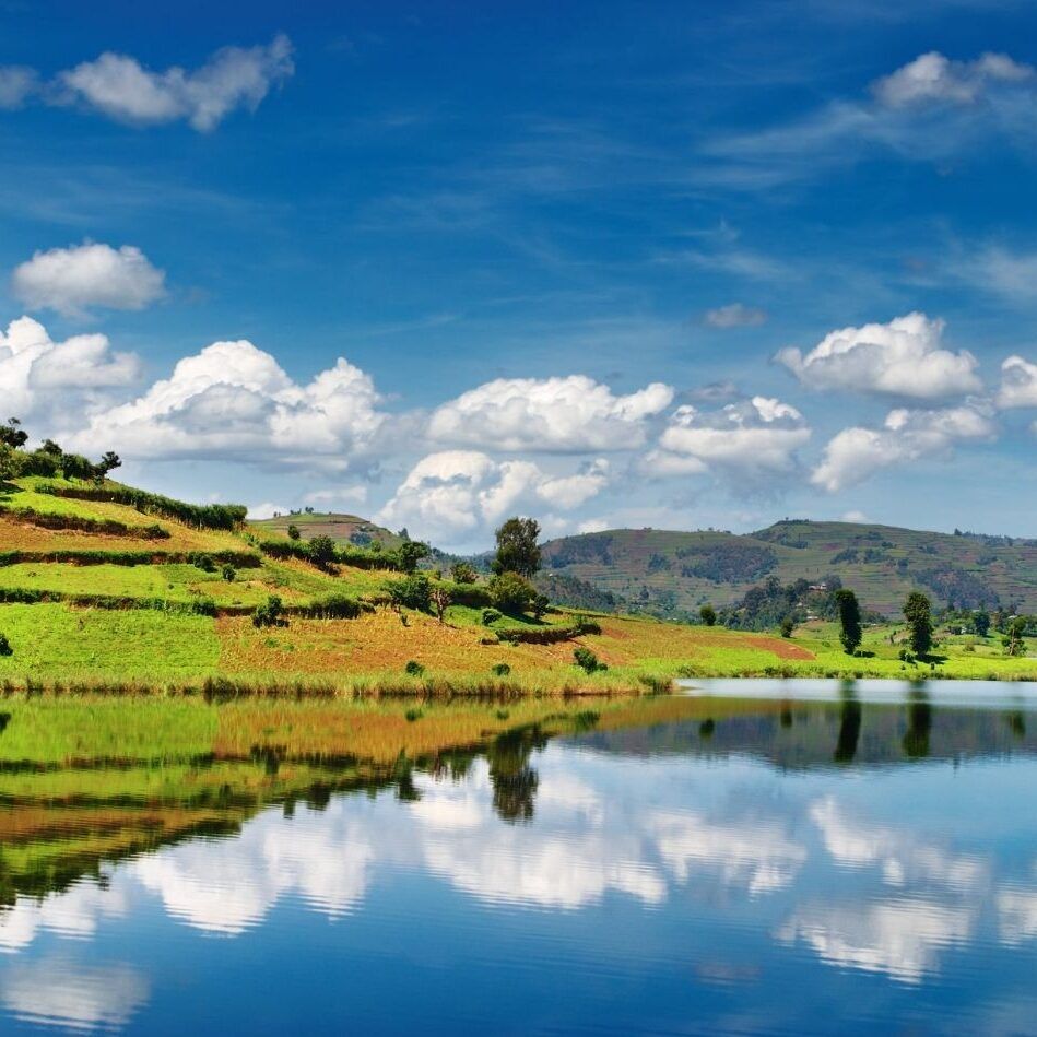 Scenic view of Lake Bunyonyi in Uganda, with rolling green hills, farmland, and fluffy clouds reflected in the calm waters of the lake.