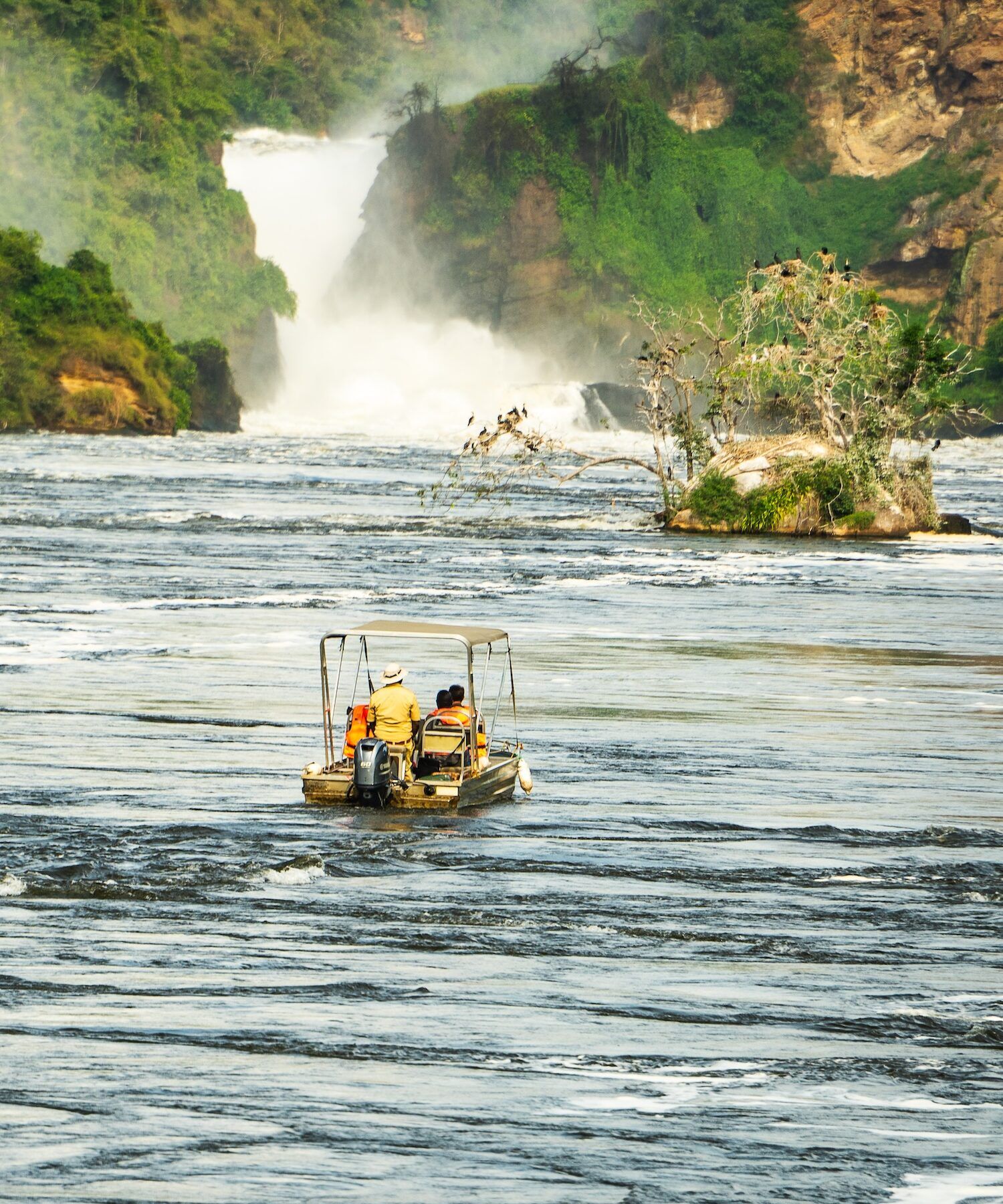 Boat safari on the Nile River approaching Murchison Falls in Uganda, where the powerful waterfall crashes through a narrow gorge surrounded by lush green vegetation.