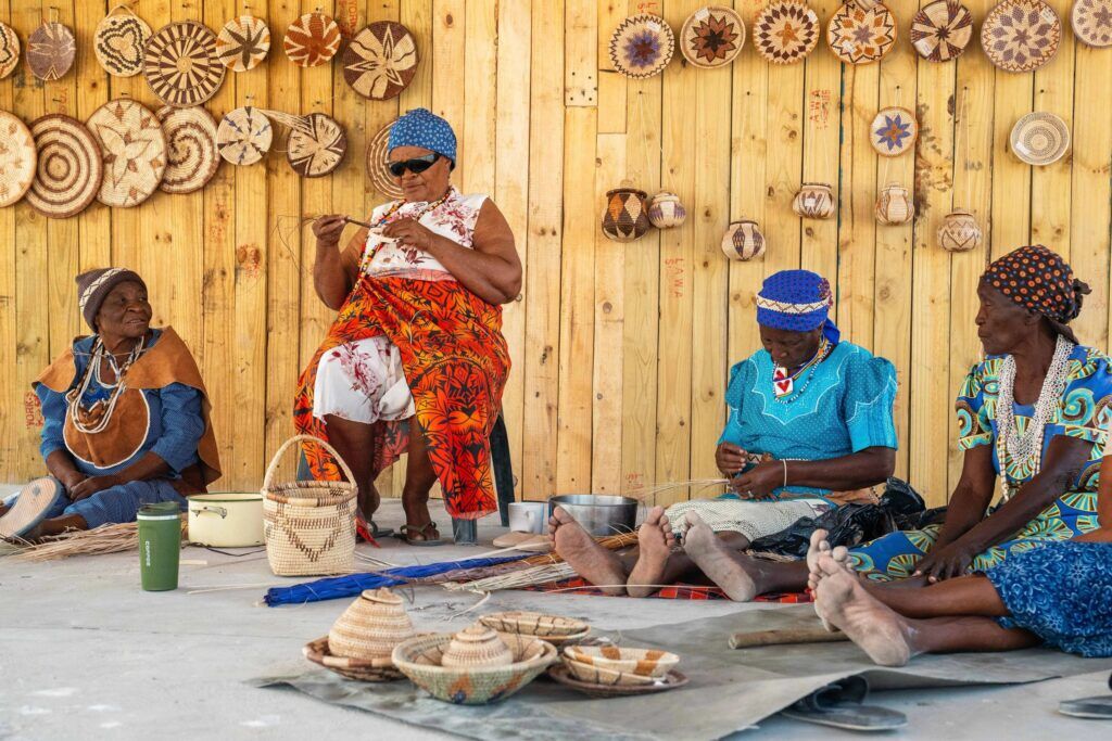 Four Khwai women sitting in a workshop surrounded by braided baskets working on arts and crafts